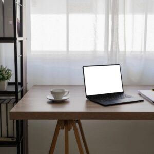 Desk with a laptop, a cup on a saucer, and a stack of papers beside a window with sheer white curtains