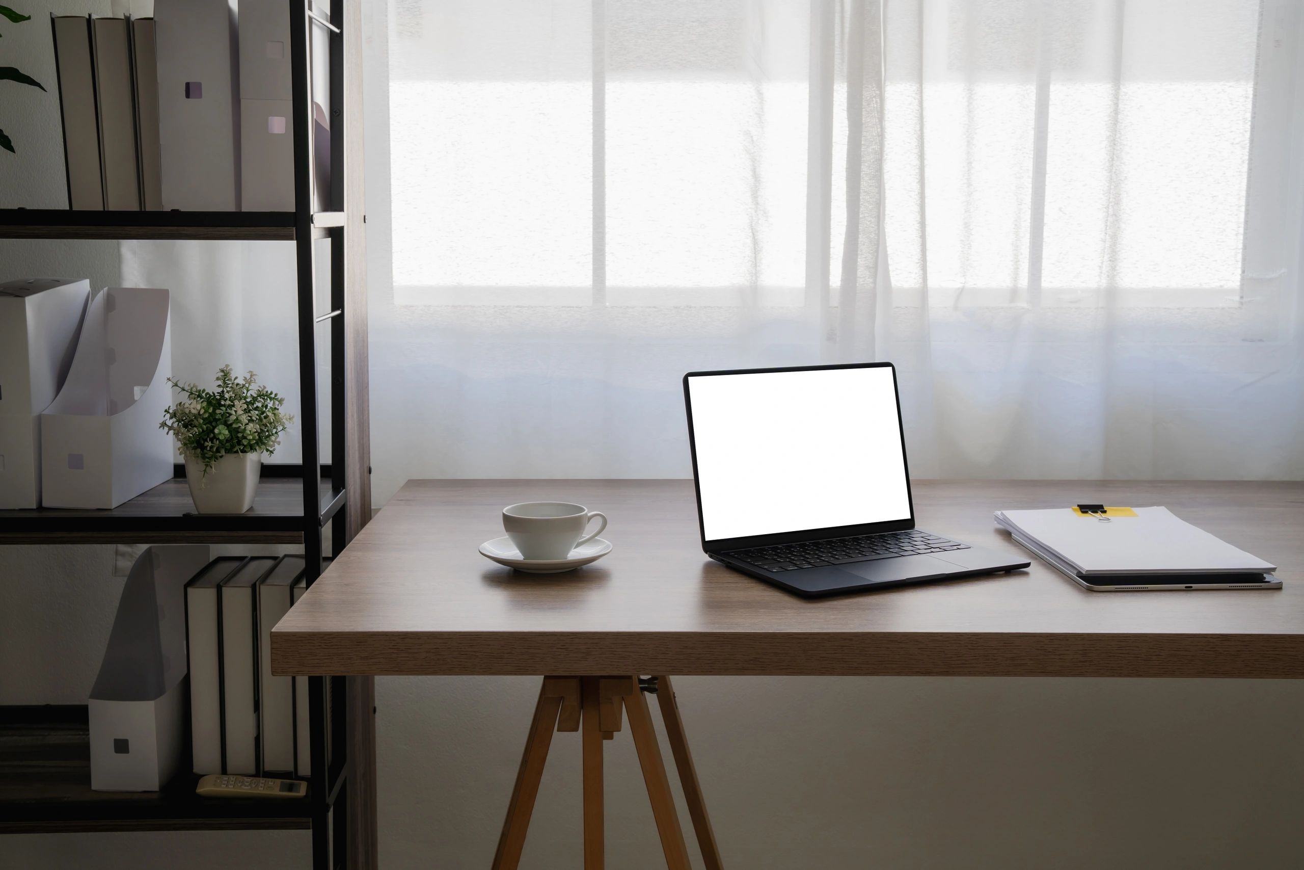 Desk with a laptop, a cup on a saucer, and a stack of papers beside a window with sheer white curtains